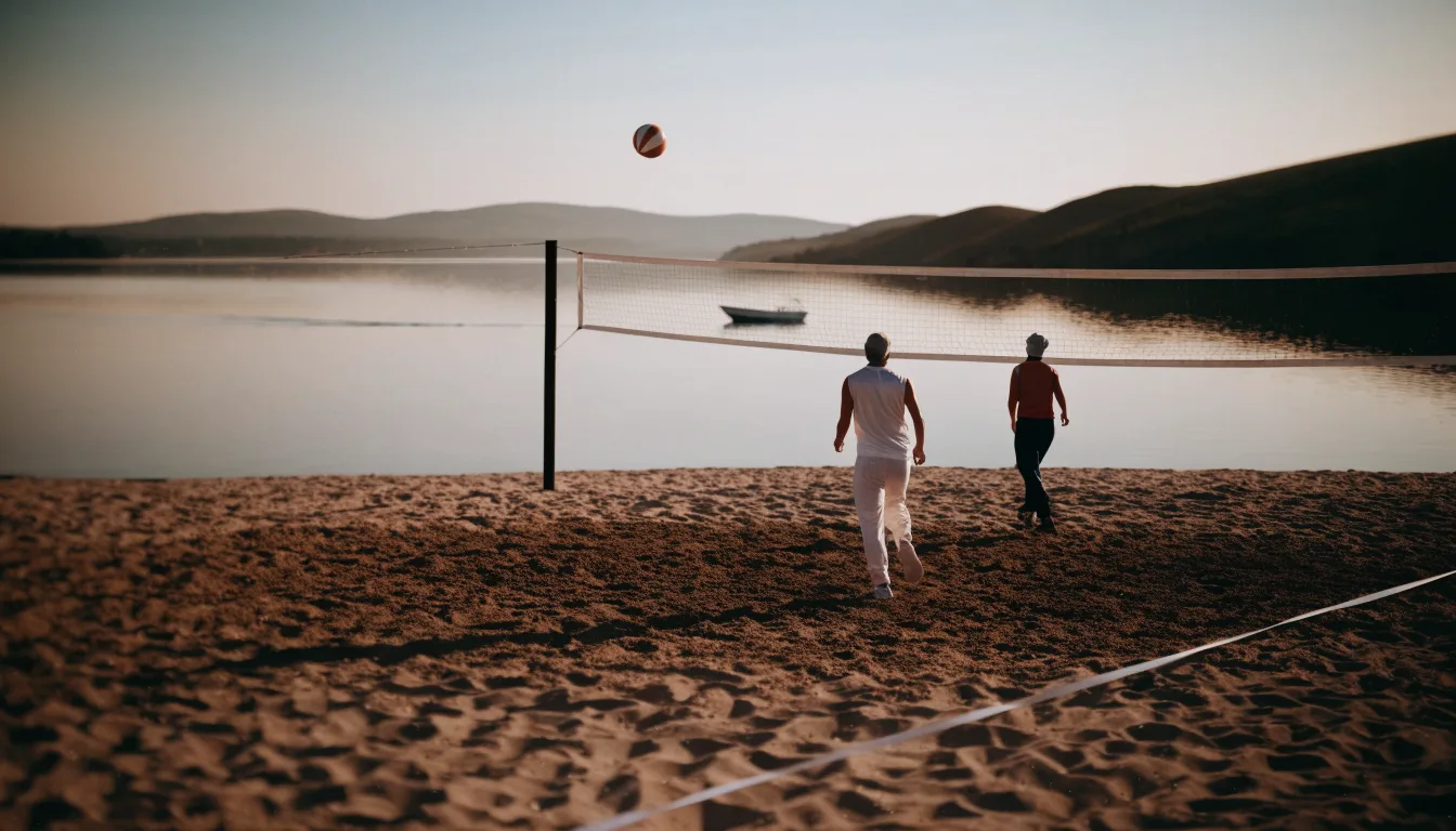 Ein Volleyball liegt im Sand eines Spielfelds am Seeufer, im Hintergrund gleitet ein Boot über das Wasser.
