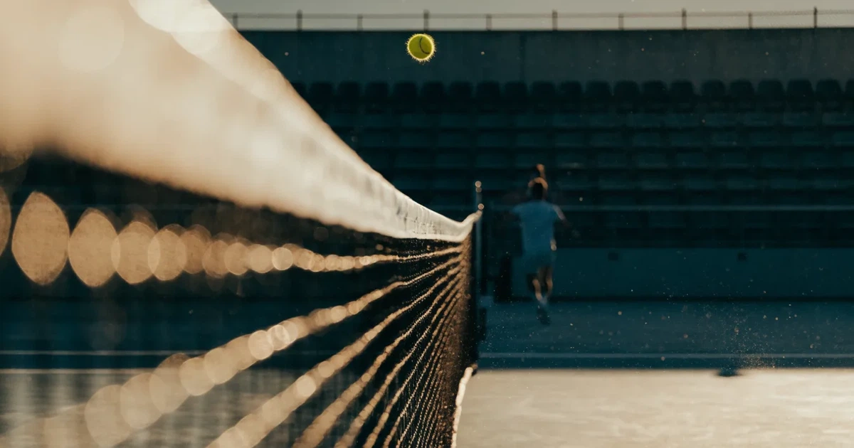 Ein Tennisnetz in Nahaufnahme im Gegenlicht der tiefstehenden Sonne, feine Staubkörner schweben über dem Platz.