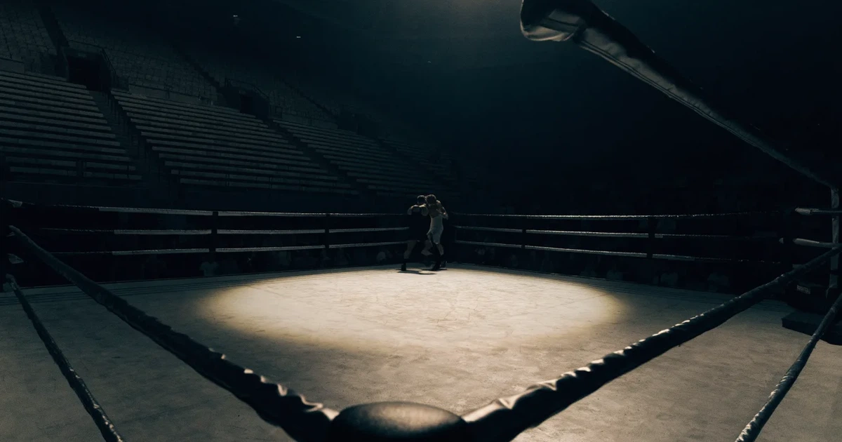 Ein leerer Boxring mit gespannten Seilen unter hellem Hallenlicht, dahinter ansteigende Zuschauerränge in einer grossen dunklen Halle