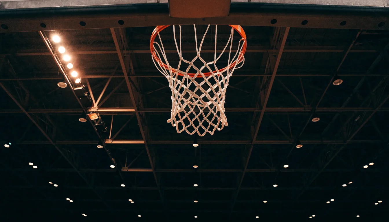 Ein Basketballkorb von unten gesehen, darüber eine Hallendecke mit Scheinwerfern und Stahlträgern.
