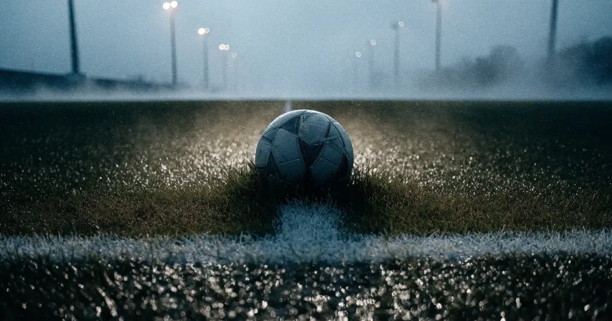Ein Fussball liegt auf der Strafraumlinie eines nassen Rasens, dahinter stehen Flutlichtmasten im aufsteigenden Nebel.
