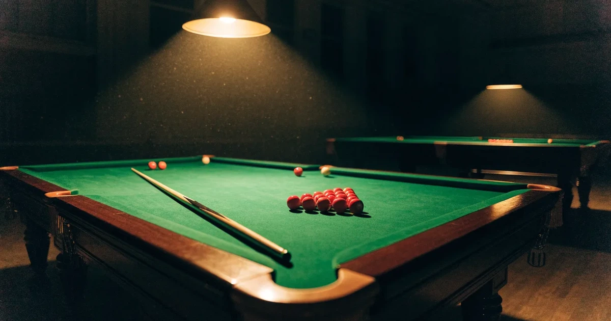 Ein Snookertisch mit grünem Tuch und einem Dreieck aus roten Kugeln unter einer tief hängenden Lampe in einer leeren Halle.