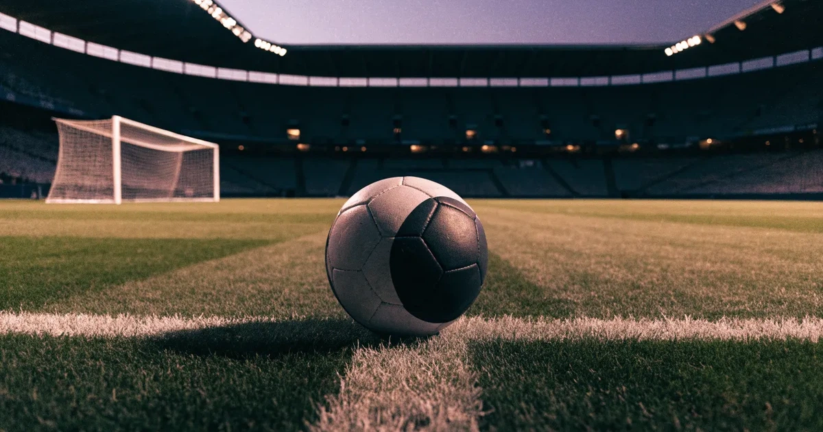 Ein Fussball liegt auf dem Anstosspunkt eines leeren Rasenplatzes, dahinter ragen die Ränge eines grossen Stadions in der Abenddämmerung auf.