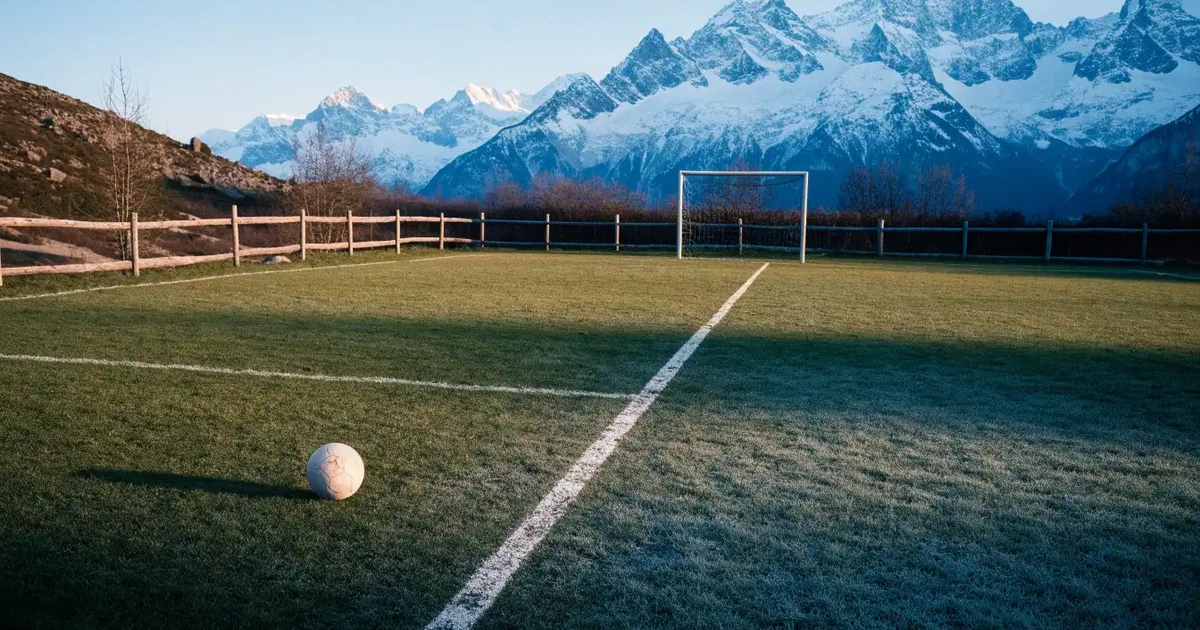 Ein Dorf-Sportplatz mit einem Ball am Strafraumrand, dahinter schneebedeckte Alpengipfel im klaren Morgenlicht