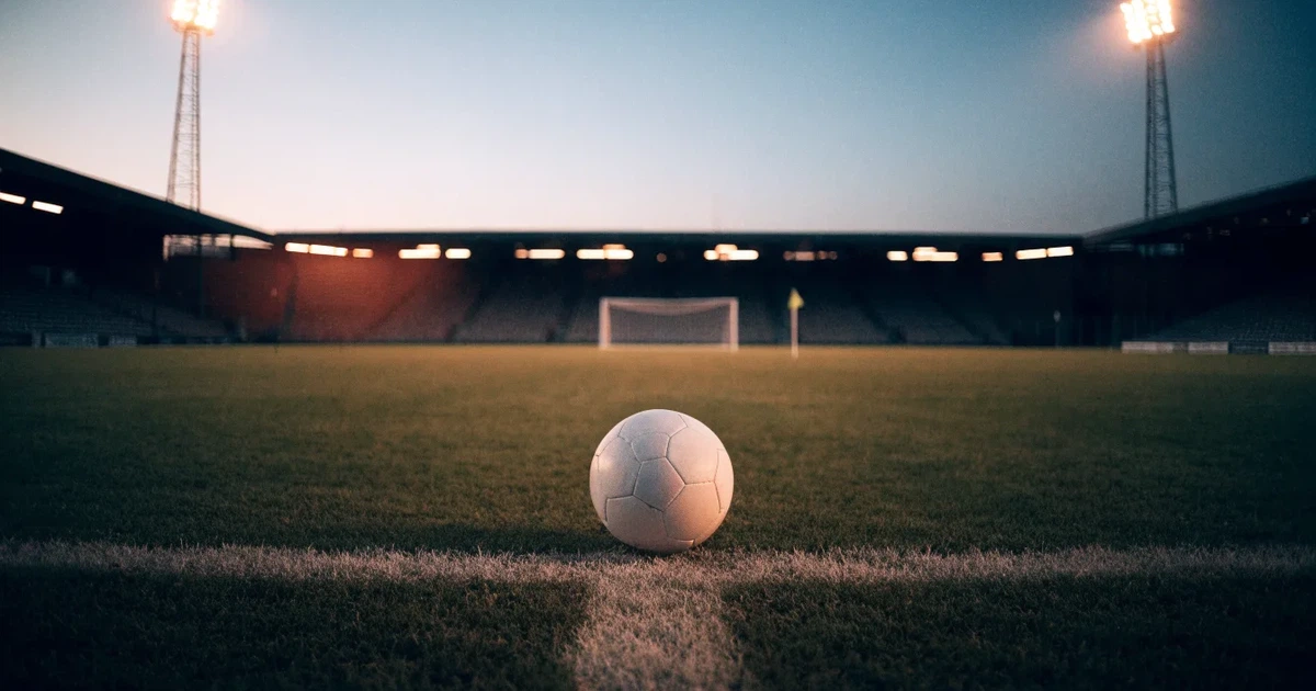 Ein Fussball liegt auf dem Anstosspunkt eines leeren Rasenplatzes in der Abenddämmerung, im Hintergrund leuchten Flutlichtmasten.